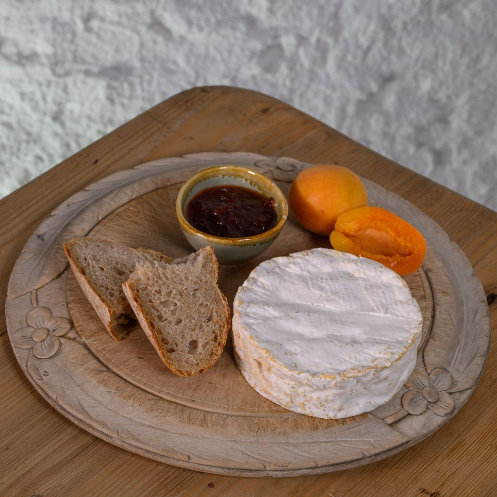 Round wooden board with a wheel of Baron Bigod cheese, bread slices, and apricots on a textured surface.