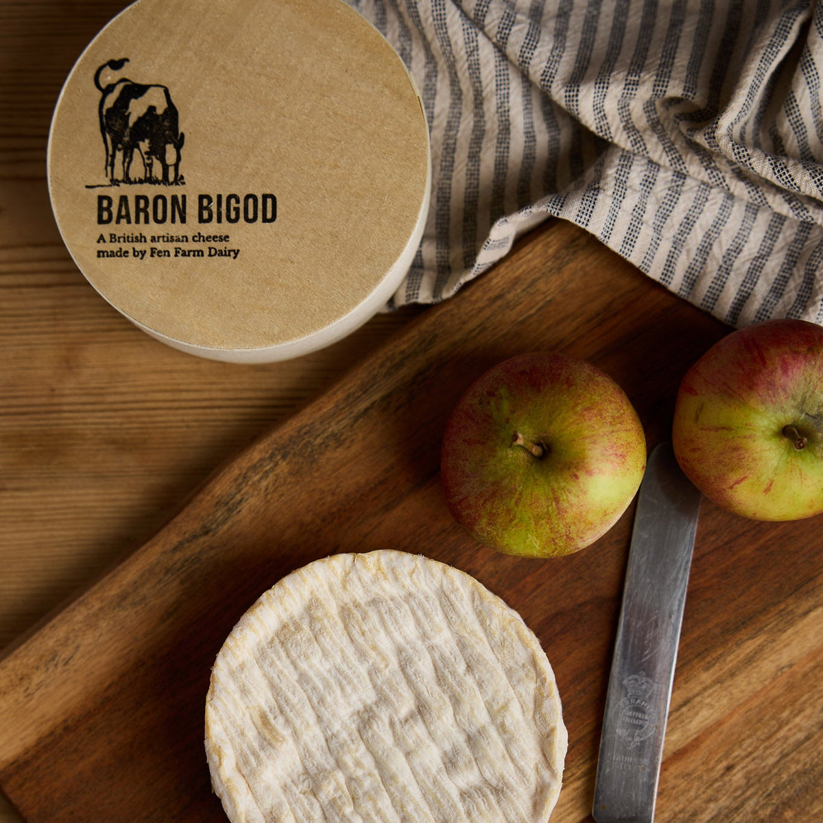 Baron Bigod cheese with its Fen Farm Dairy box, apples and a cheese knife on a wooden board.