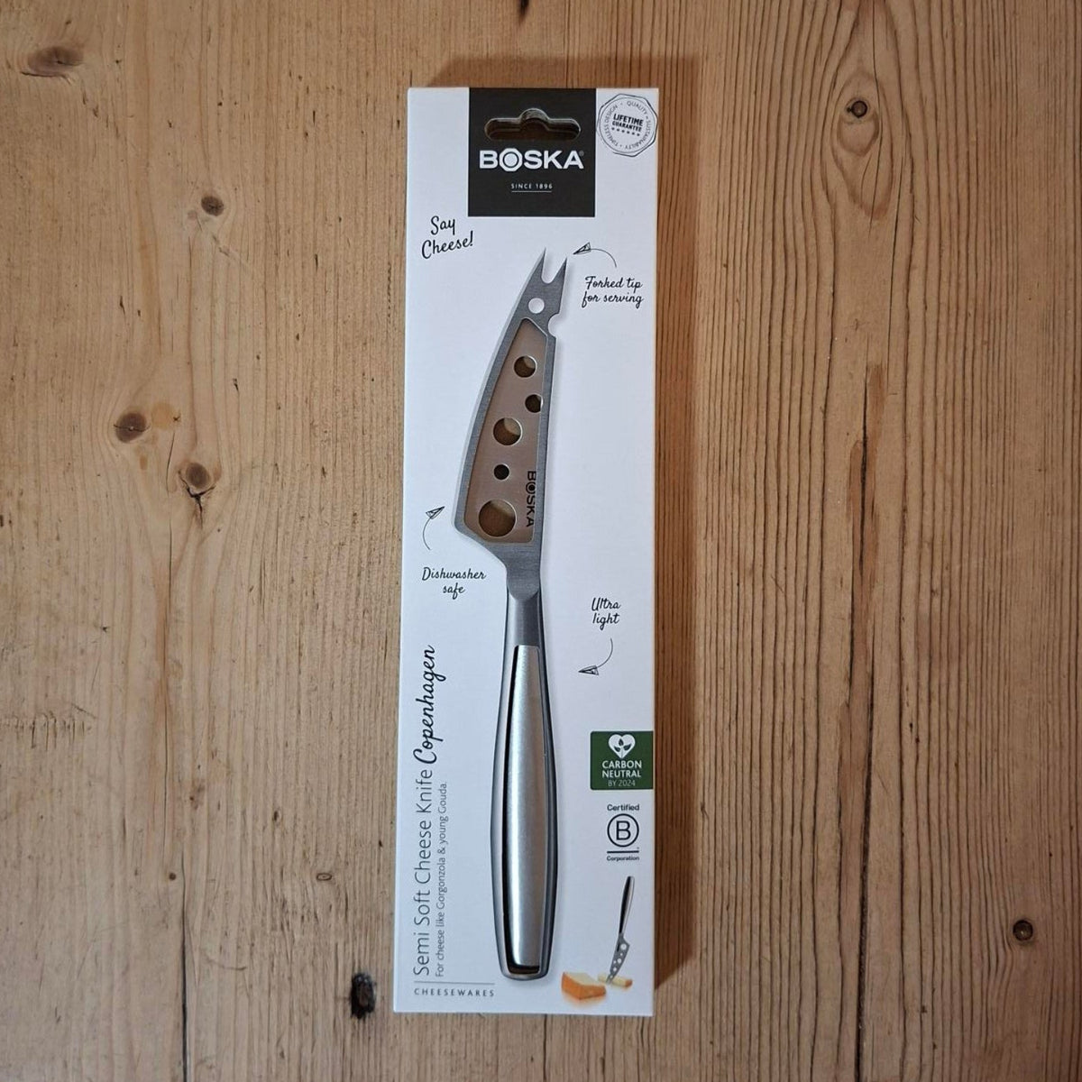A Boska cheese knife in its original white packaging, displayed vertically on a light brown wooden surface.
