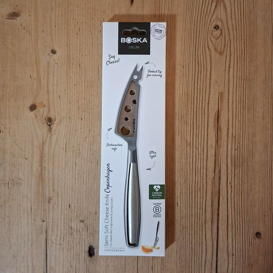 A Boska cheese knife in its original white packaging, displayed vertically on a light brown wooden surface.