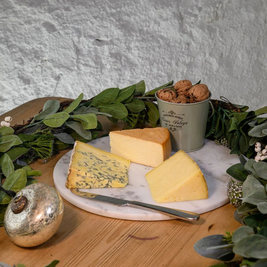 Three types of cheese on a marble board with a small container of nuts and greenery in the background.