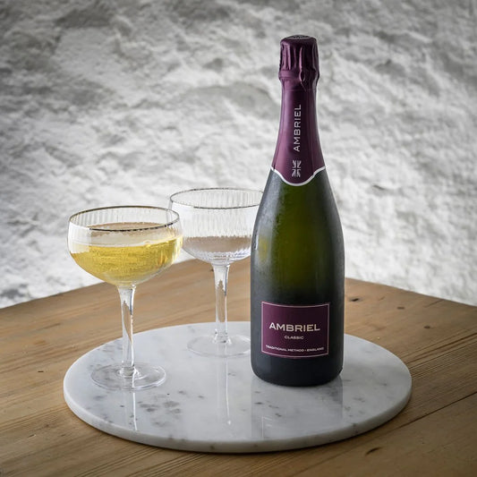 Bottle of Ambriel English Sparkling with two glasses on a marble coaster against a textured wall.