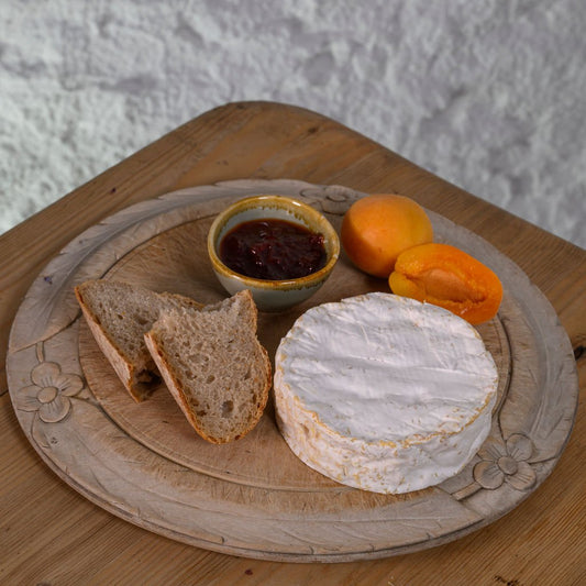 Round wooden board with a wheel of Baron Bigod cheese, bread slices, and apricots on a textured surface.