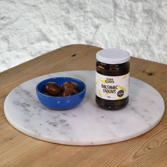 Jar of balsamic onions and a bowl of onions on a marble board with a wooden surface.