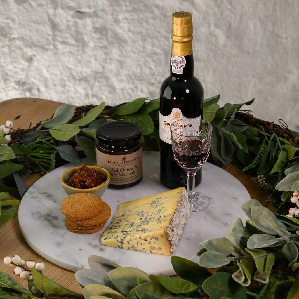 Bottle of Graham's port wine with a glass, stilton cheese, crackers, and a jar on a marble board with greenery.