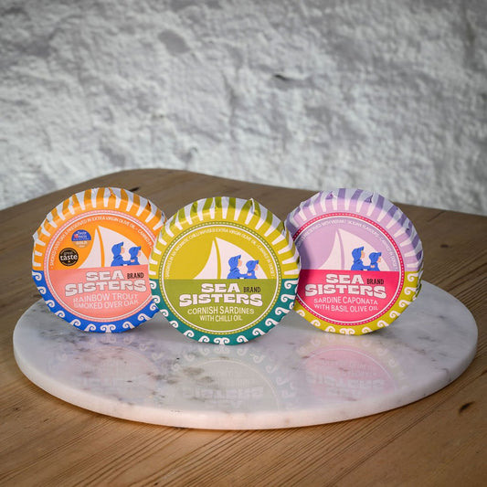 Three colorful tins of Sea Sisters tinned fish, on a marble coaster with a wooden surface and gray stone background.