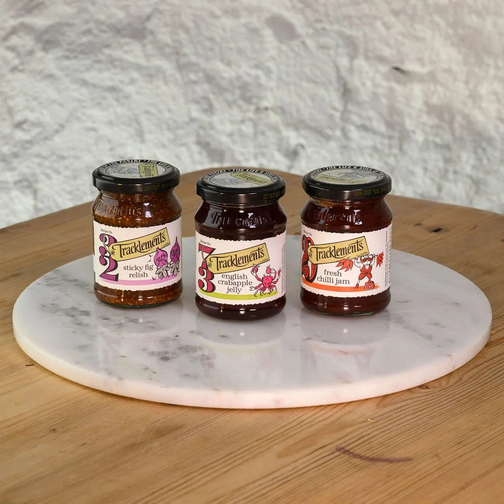 Three jars of Tracklements preserves on a marble platter with a wooden surface and marble wall background.