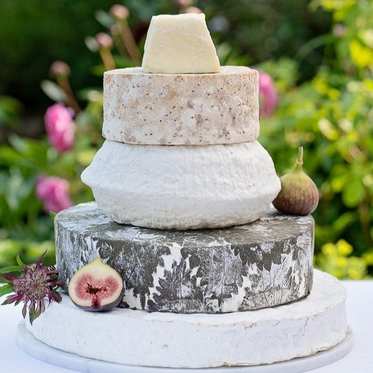 Three stacked rustic cheese wheels on a white pedestal plate, styled with figs and a flower in a blurred garden setting.